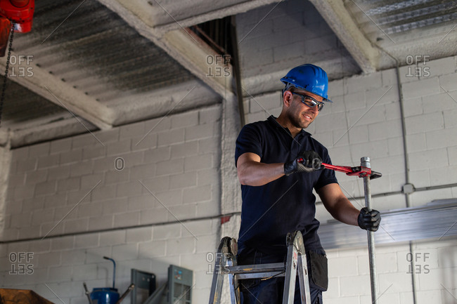 Low angle of cheerful male worker in hardhat and protective glasses standing on ladder and installing metal pipe with plumber wrench while working in workshop