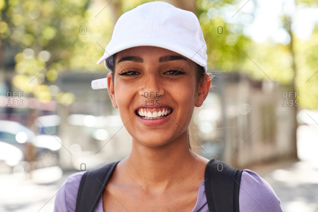 Optimistic female adolescent in casual outfit and white cap with backpack smiling and looking at camera while standing against blurred urban street in summer day