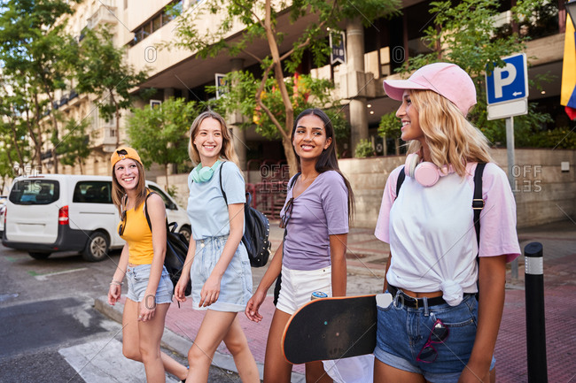 Group of cheerful multiracial teen girls in casual clothes with headphones and skateboard having fun and chatting happily while walking together on city street in summertime