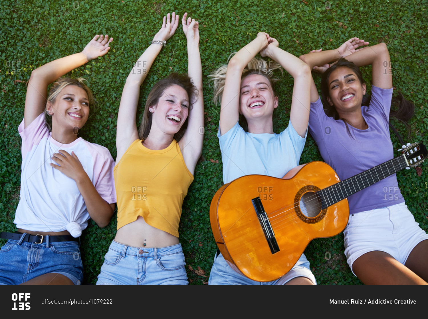 From above of group of content diverse teen girlfriends in\
casual outfits with guitar chilling together on green grass and\
enjoying summer holidays in park stock photo - OFFSET