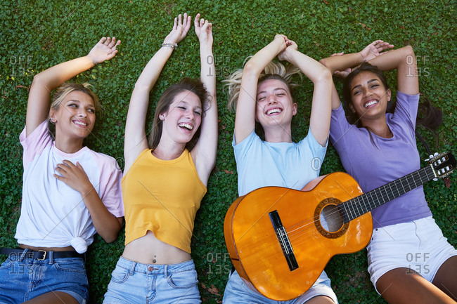 From above of group of content diverse teen girlfriends in casual outfits with guitar chilling together on green grass and enjoying summer holidays in park