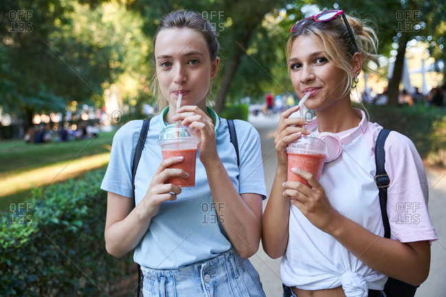 Joyful blonde teen female friends with fruit popsicle and cold drink chatting happily while spending summer day together in park