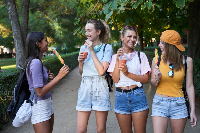 Group of cheerful diverse hipster girlfriends enjoying takeaway cold beverages and fruit ice cream in summer park