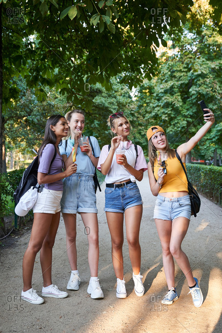 Group of cheerful diverse hipster girlfriends taking selfie on smartphone while enjoying takeaway cold beverages and fruit ice cream in summer park