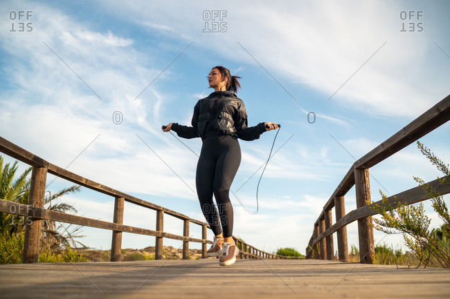 From below full body of fit sporty female in black sportive outfit jumping with skipping rope on wooden walkway during outdoor fitness training in summer day