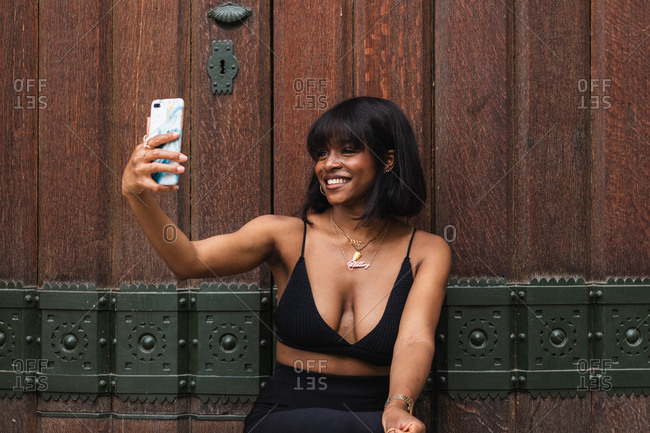 Smiling young African American female wearing black top with decollete photographing selfie on cellphone