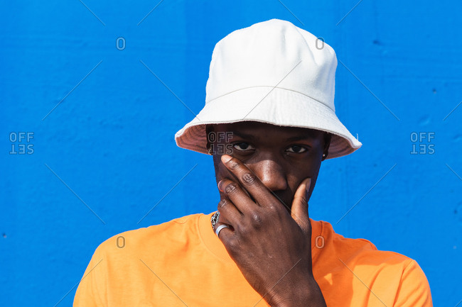 Young African American male in bright yellow t shirt and white cap touching face with hand while standing against blue wall