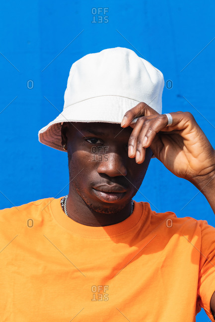 Young African American male in bright yellow t shirt adjusting white cap while standing against blue wall