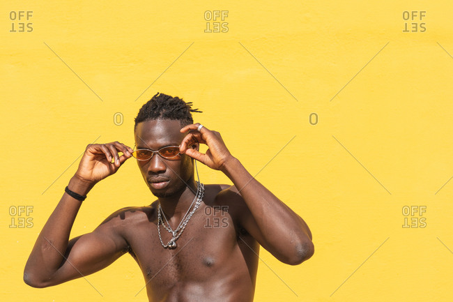 Serious young shirtless African American male in stylish sunglasses and with metal necklaces posing against yellow wall with striped shadow