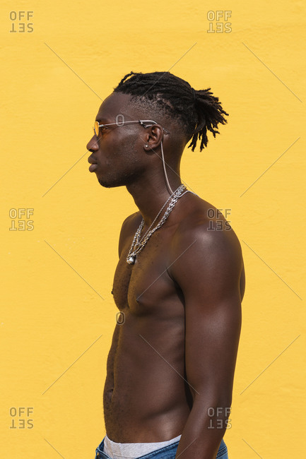 Serious young shirtless African American male in stylish sunglasses and with metal necklaces posing against yellow wall with striped shadow