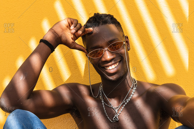 Positive young shirtless African American male in stylish sunglasses and with metal necklaces sitting against yellow wall with striped shadow