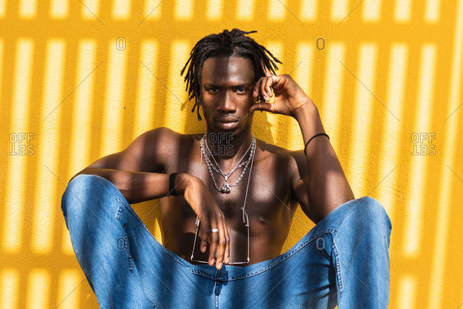 Serious young shirtless African American male in stylish sunglasses and with metal necklaces sitting against yellow wall with striped shadow