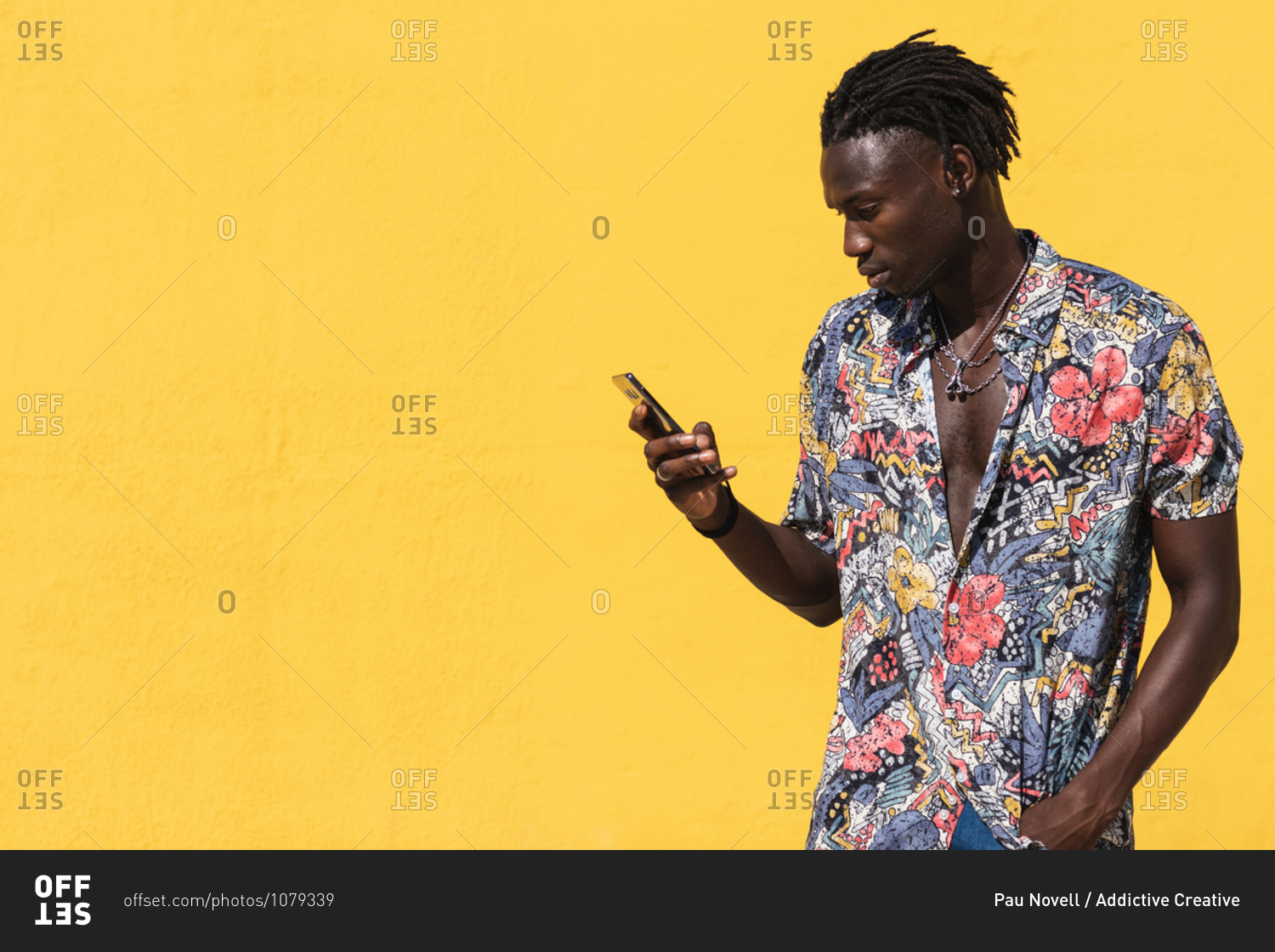 Confident young hipster African American male with dreadlocks wearing trendy summer shirt with floral print and jeans using mobile phone against yellow background