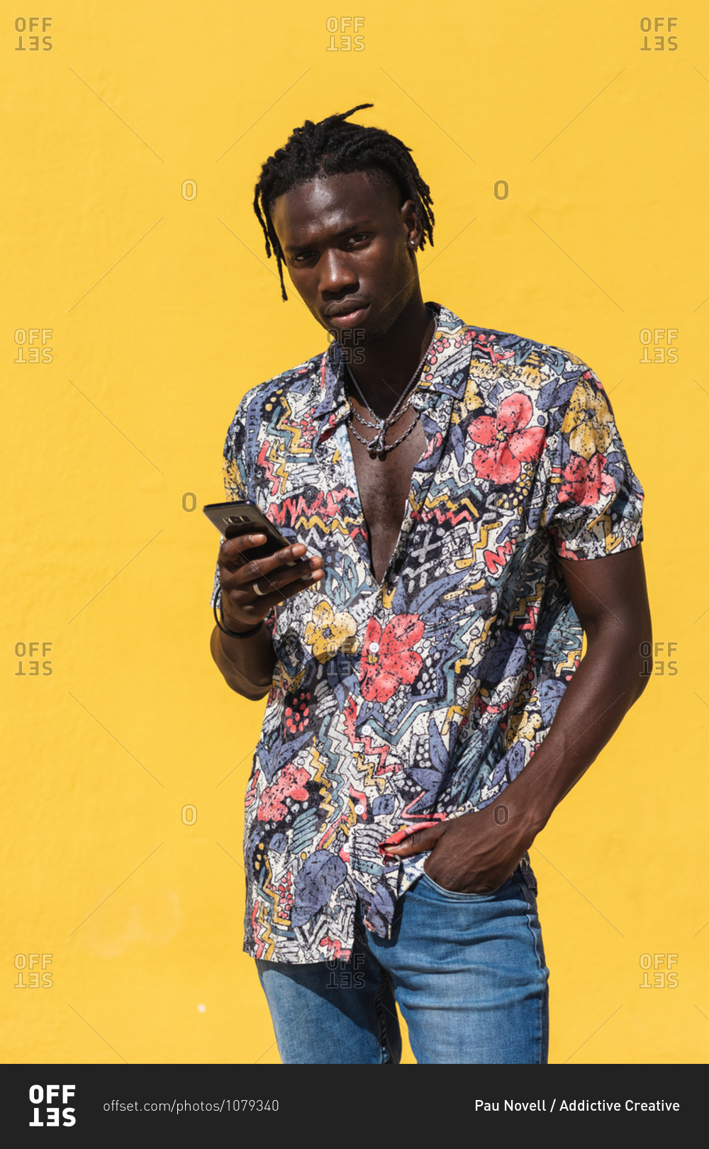 Confident young hipster African American male with dreadlocks wearing trendy summer shirt with floral print and jeans using mobile phone and looking at camera against yellow background