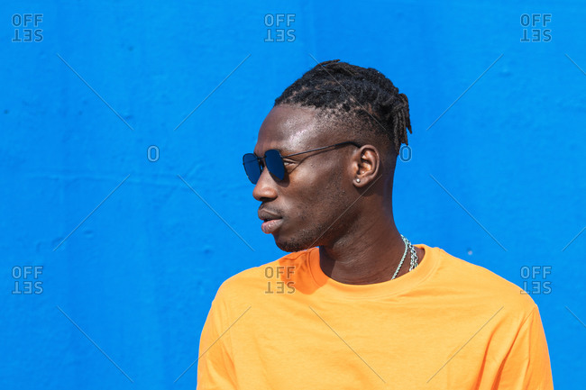 Serious young African American guy in bright yellow t shirt and trendy sunglasses looking away against blue background