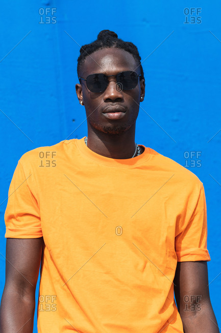 Serious young African American guy in bright yellow t shirt and trendy sunglasses looking at camera against blue background