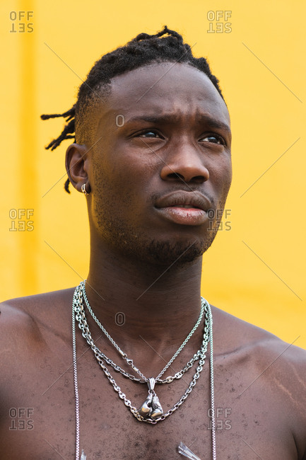 Serious young African American guy with naked torso dreadlocks and metal chains against yellow background looking away