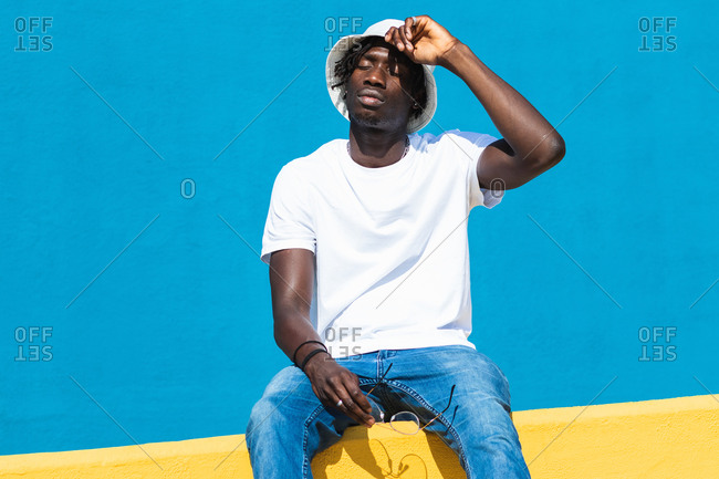 Low angle full body of relaxed young black male in casual outfit with panama hat and sunglasses siting on bright colorful fence and enjoying sunny summer day