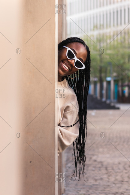 Happy young black female in stylish sunglasses peeking from behind pillar and smiling on city street