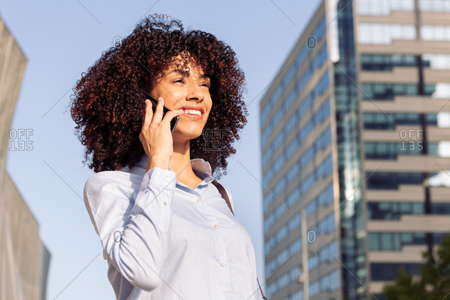 From below of cheerful ethnic female entrepreneur standing in downtown and having conversation on mobile phone while looking away