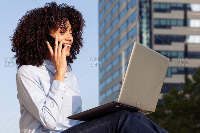 Side view of focused happy female entrepreneur sitting on stone bench in park and working remotely while speaking on smartphone and using laptop