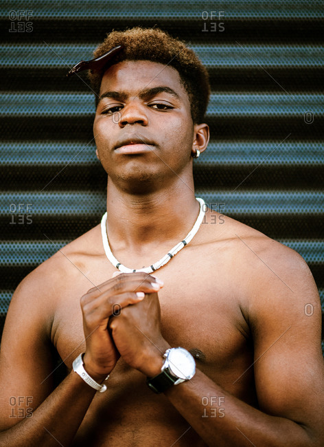 Confident African American male with naked torso and in various luxury necklaces standing in city and looking at camera