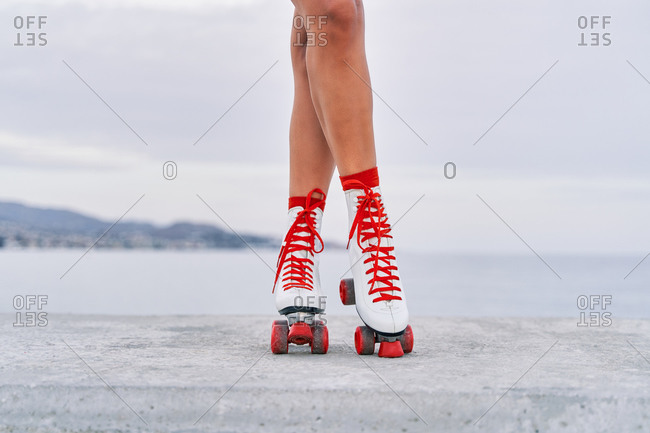 Crop faceless female with slim legs and in red and white roller skates standing on stone border on background of sea