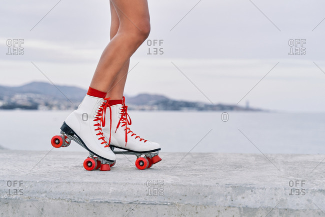 Crop faceless female with slim legs and in red and white roller skates standing on stone border on background of sea