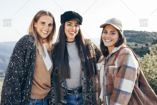 Company of positive female friends in stylish casual clothes cuddling together while standing on hill in mountains