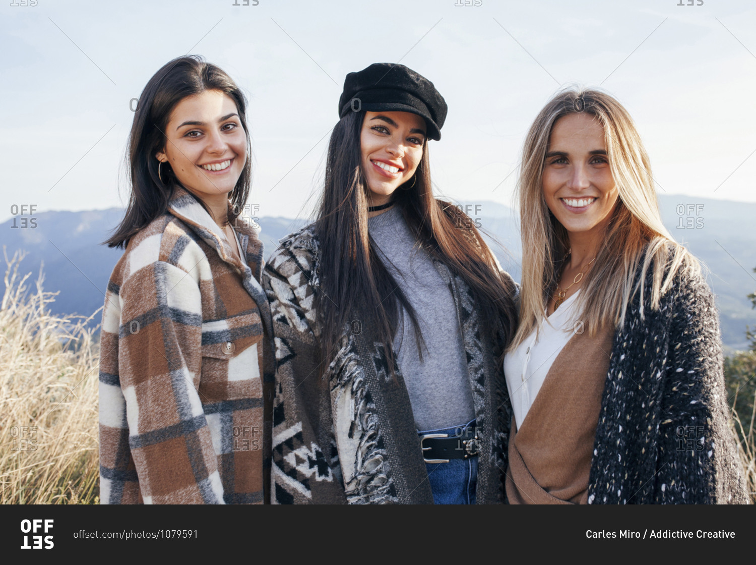 Company of positive female friends in stylish casual clothes cuddling together while standing on hill in mountains