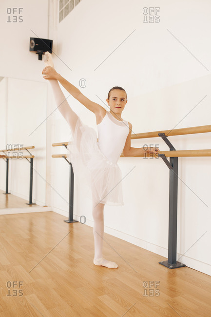 Charming teenage ballerina in pointe shoes stretching legs near ballet barre in dance hall