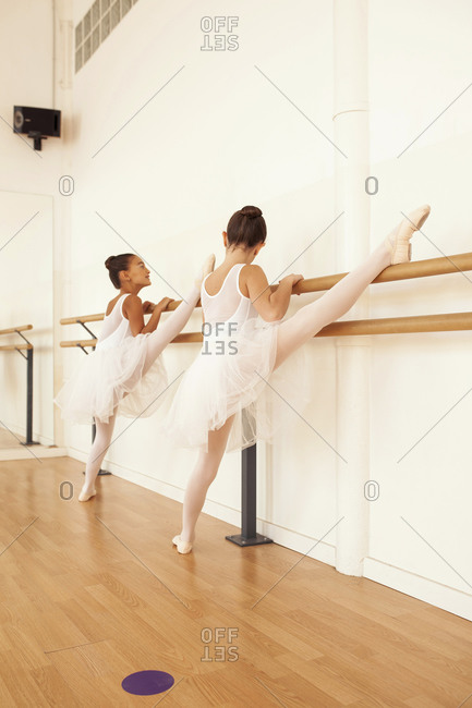 Low angle of charming teenage ballerinas in pointe shoes stretching legs near ballet barre in dance hall