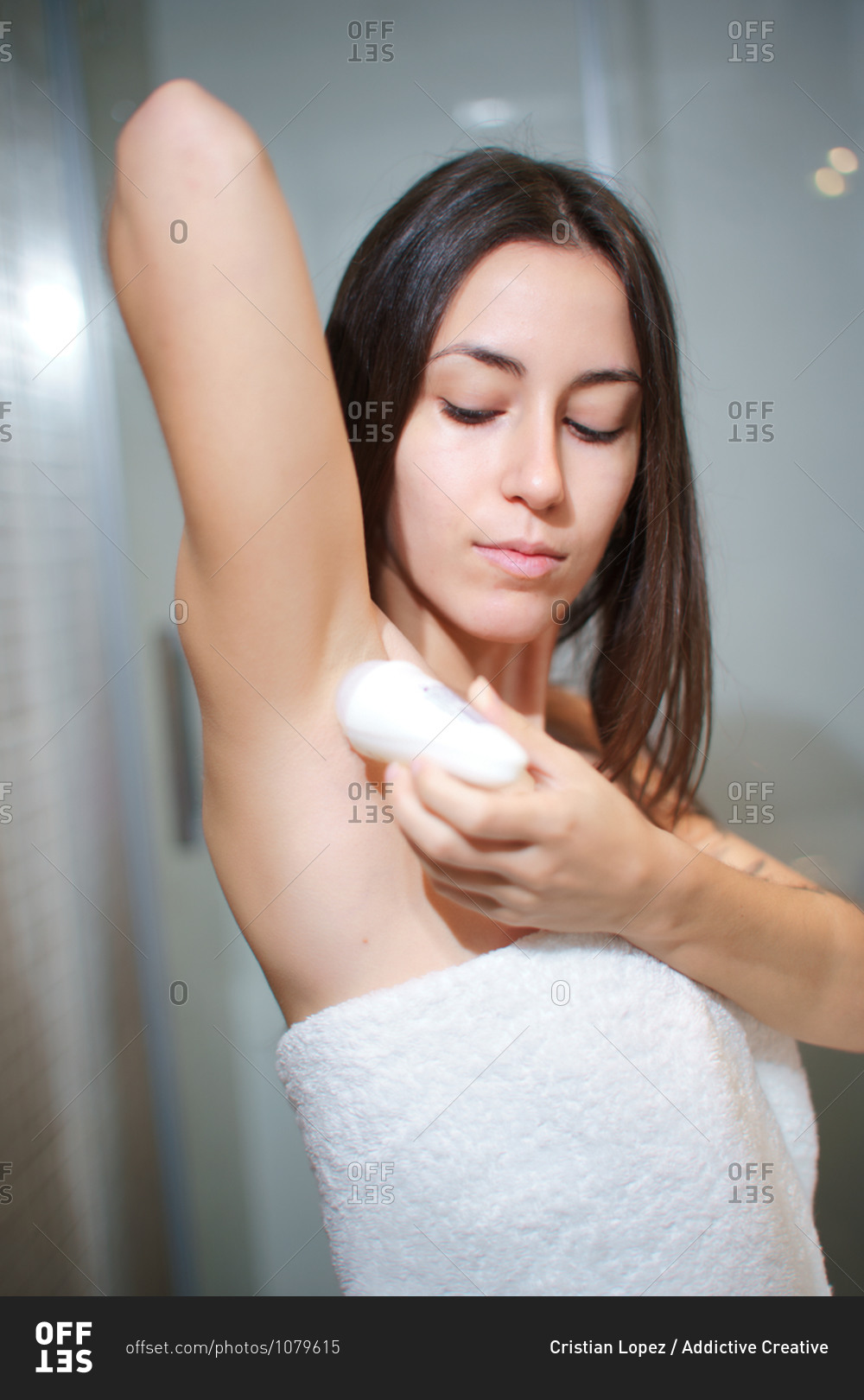 Side view of delicate female in towel standing in bathroom and applying antiperspirant on armpit after taking shower