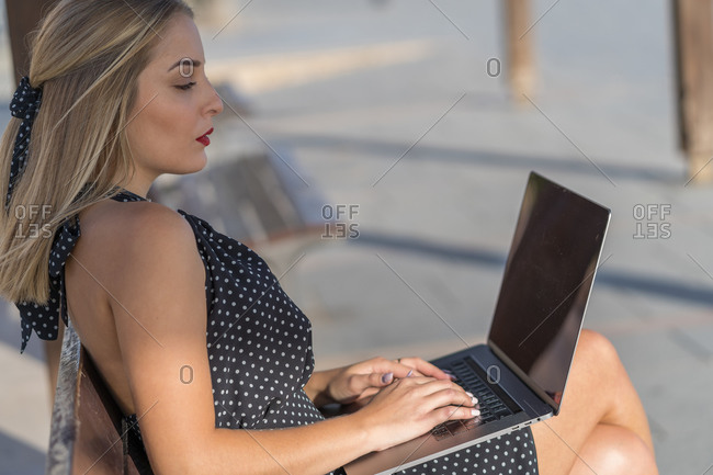 Side view of self employed woman sitting on bench on embankment and working on project while tying on laptop