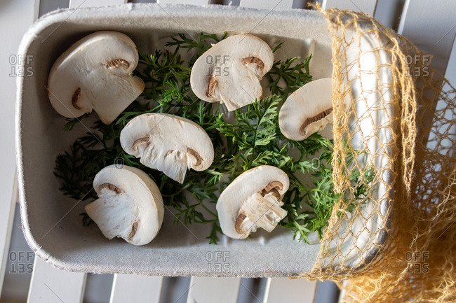 Top view of halved fresh bio organic champignons with green herbs in pot with eco sac placed on white plank surface