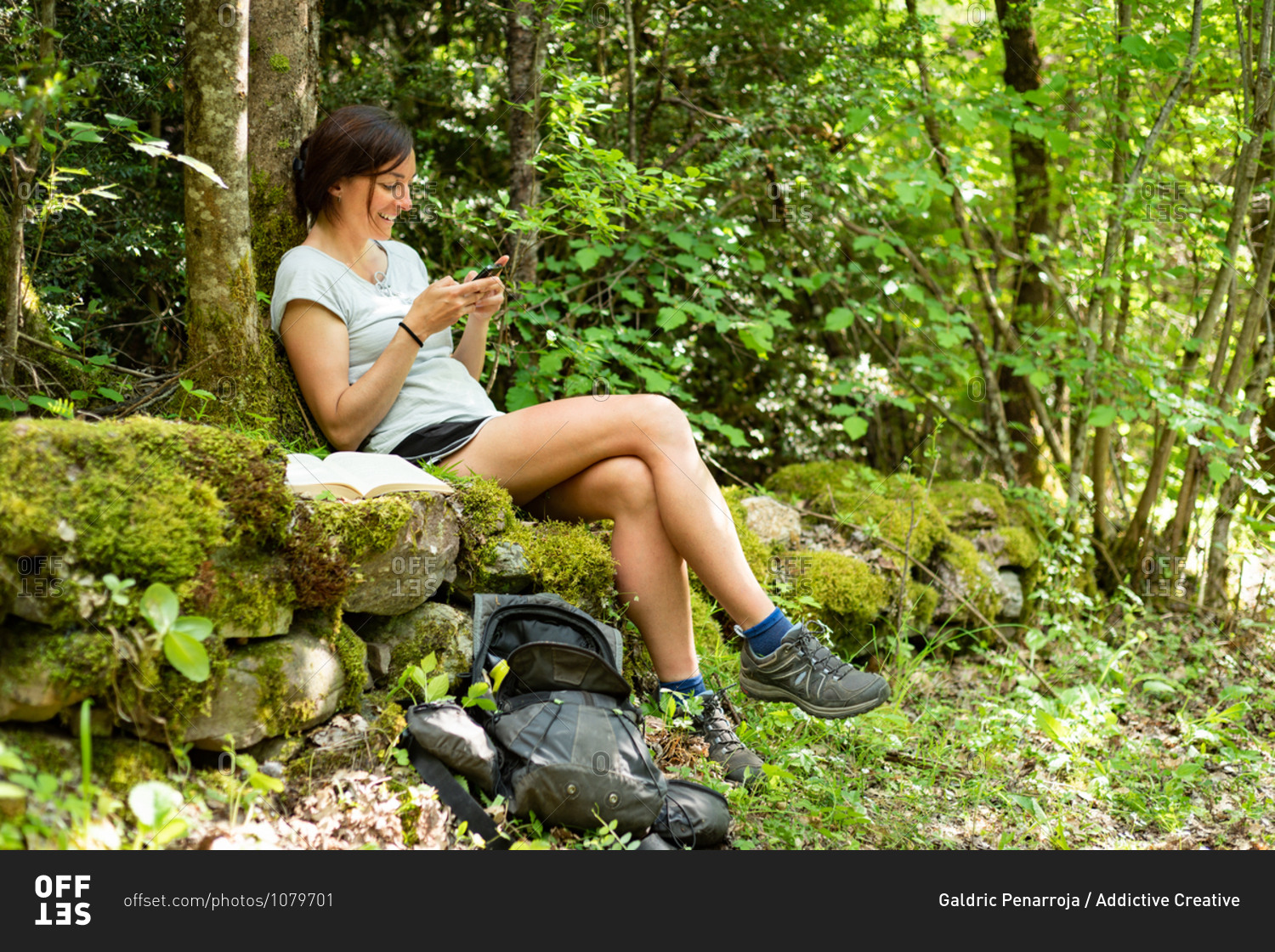 Side view of delighted female traveler relaxing on mossy stones in green woods and browsing cellphone during summer adventure