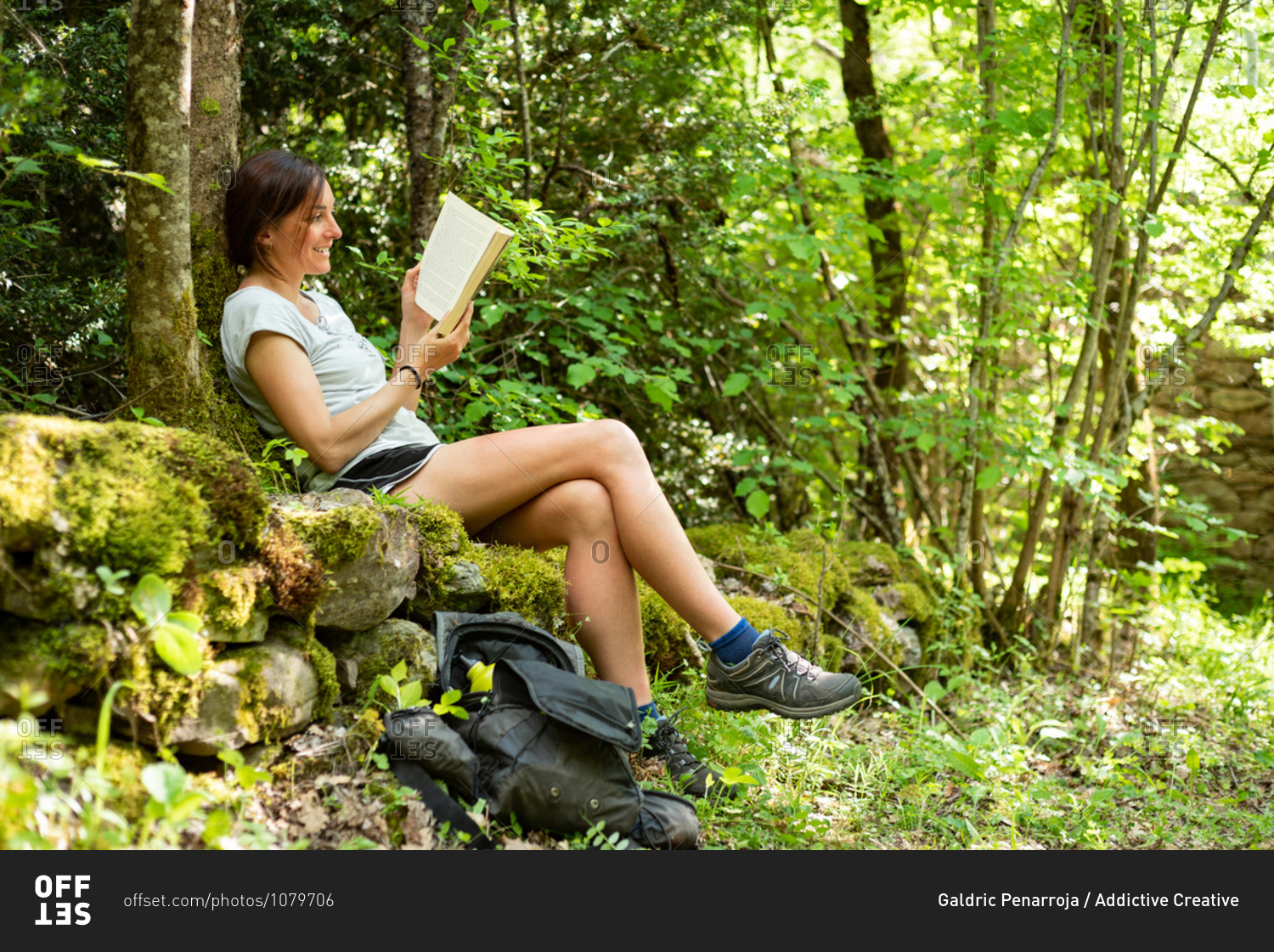 Side view of focused female sitting on mossy stones in woods and enjoying interesting story in book while spending time in nature
