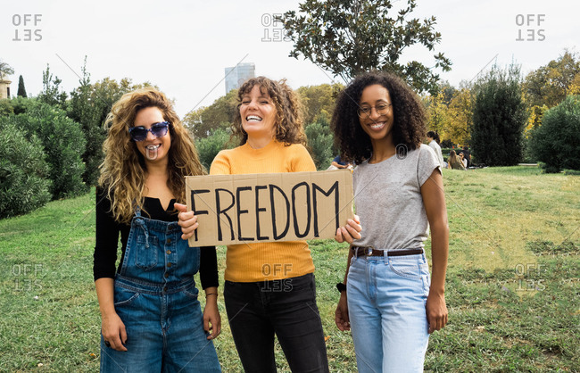 Low angle of company of multiracial female friends standing in park with poster Freedom and looking at camera