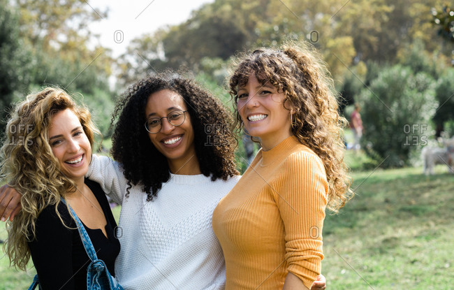 Group of young multiracial female friends smiling and cuddling in park while looking at each other