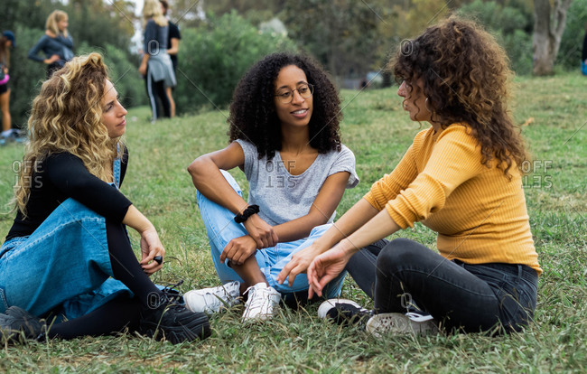 Company of diverse female friends sitting on lawn in summer park and talking to each other