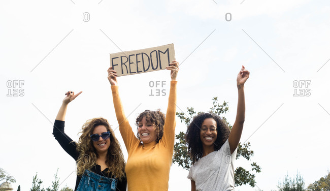 Low angle of company of multiracial female friends standing in park with poster Freedom and looking at camera