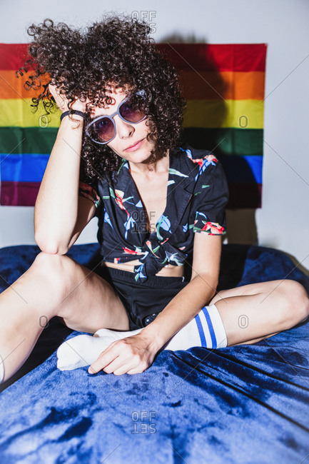 Confident female sitting on bed leaning on hand looking at camera against rainbow flag