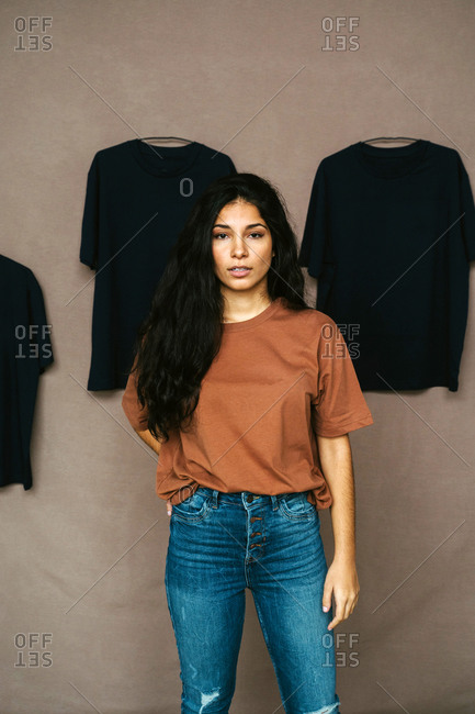 Ethnic female in casual outfit standing in studio with t shirts on wall and looking at camera