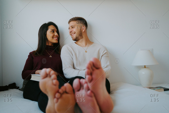 Smiling barefoot boyfriend and girlfriend sitting on comfortable bed and enjoying weekend while looking at each other