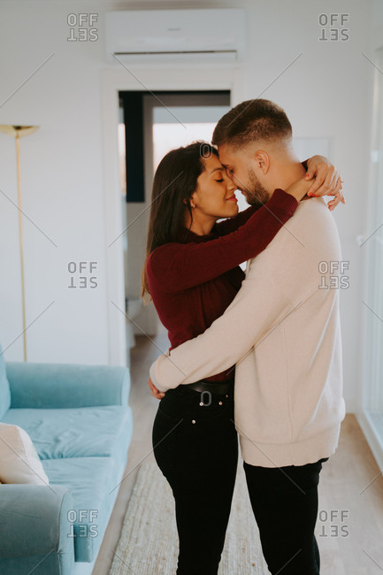 Side view of couple in love standing in living room and cuddling while touching faces with closed eyes