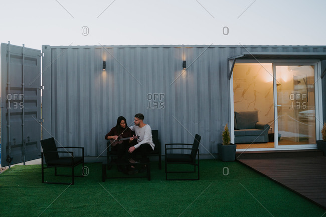 Tranquil couple sitting on backyard of container house and playing ukulele while enjoying music in evening
