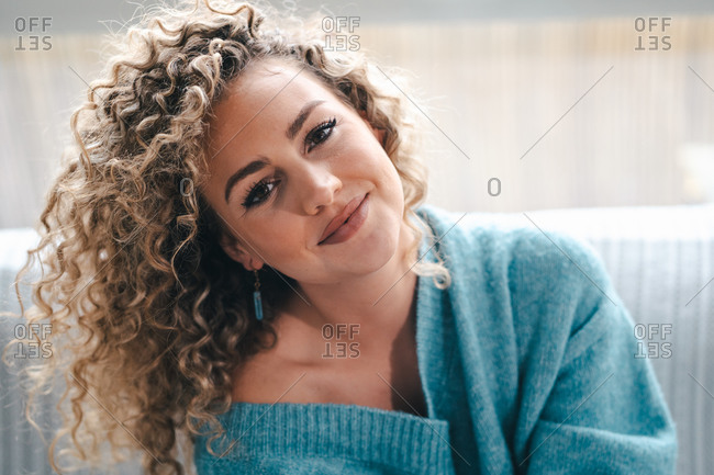 Coquette female with curly hair and in warm knitted sweater resting on comfortable sofa and looking at camera