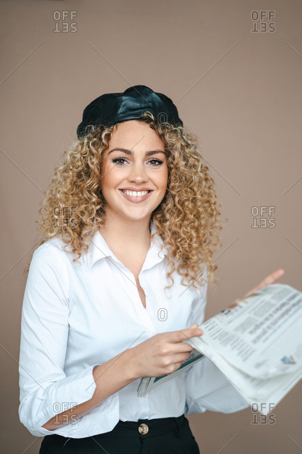 Smiling female wearing trendy beret standing in studio with newspaper and looking at camera