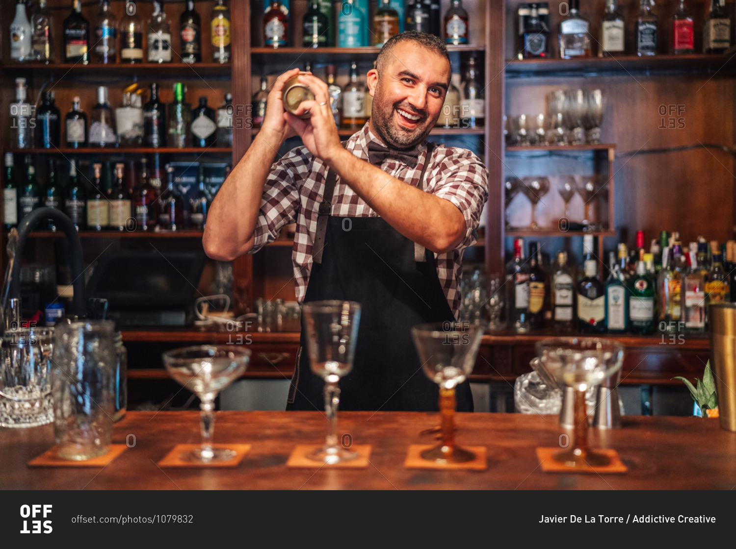 Excited middle aged mixologist in checkered shirt and apron looking at camera and mixing alcohol drink in shaker while standing behind counter with glasses in bar