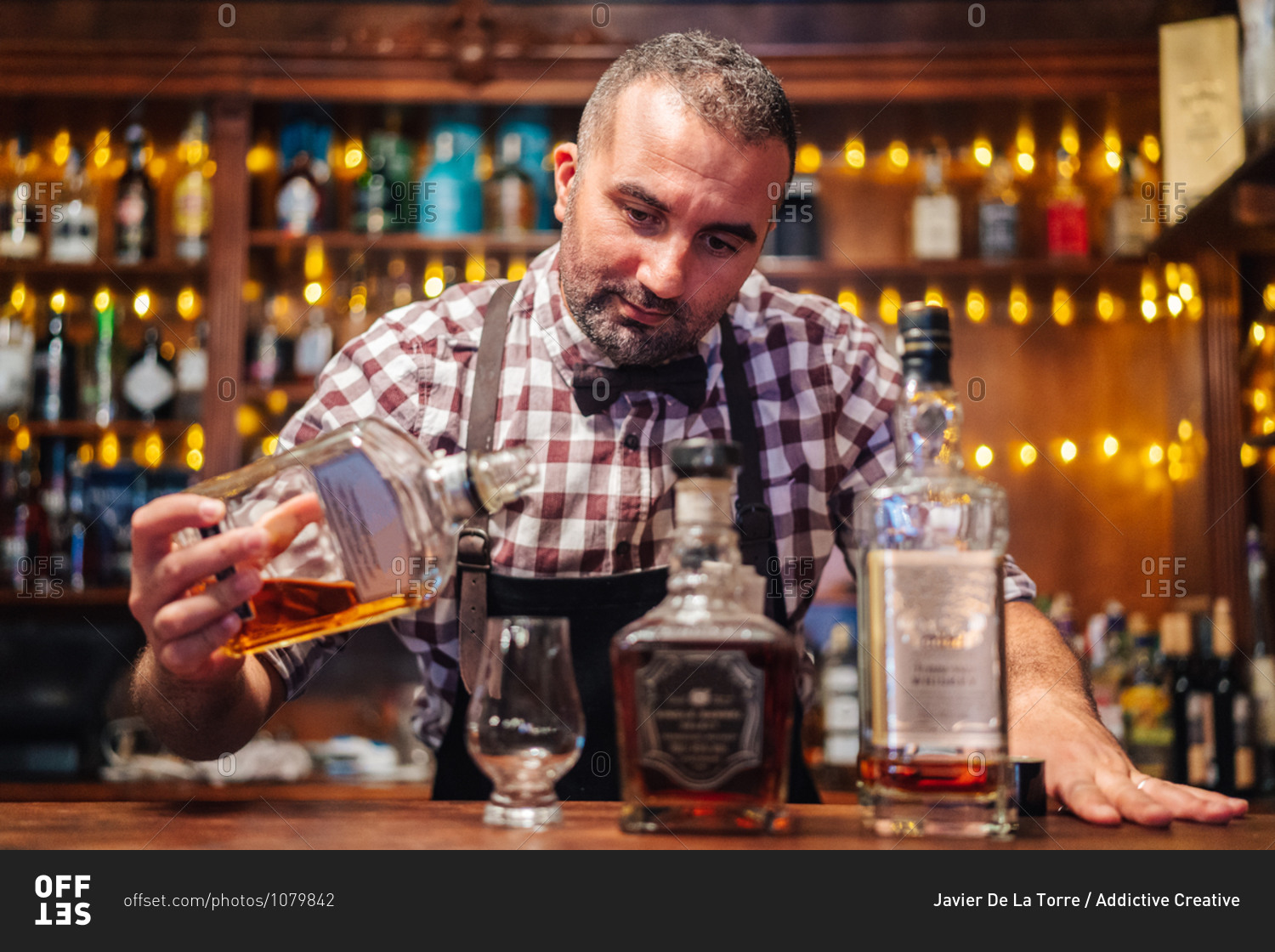 Crop brutal male bartender in apron pouring brandy in glass placed on counter with bottles of alcoholic drinks